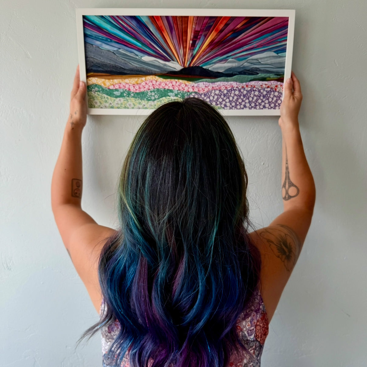 A woman with colorful hair holding a framed abstract kinusaiga art piece against a white wall.